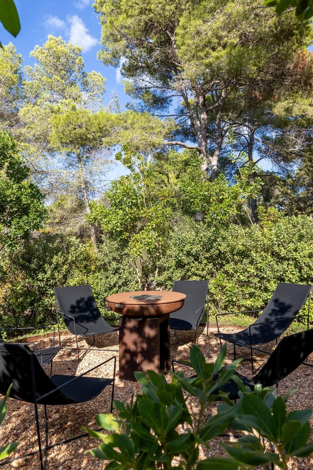 Corten steel fire pit ringed by black sling chairs in a clearing of pines and Mediterranean shrubs