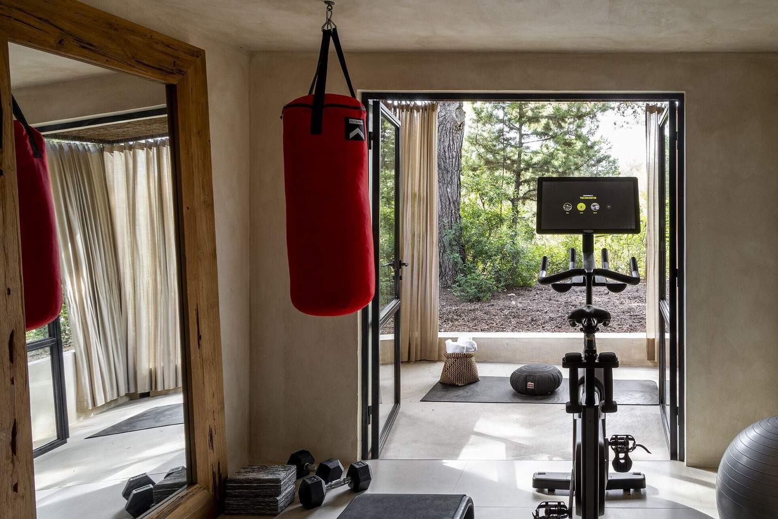 Lime-washed gym with red Outshock heavy bag, Technogym Skillbike and dumbbells, doors open to a pine grove