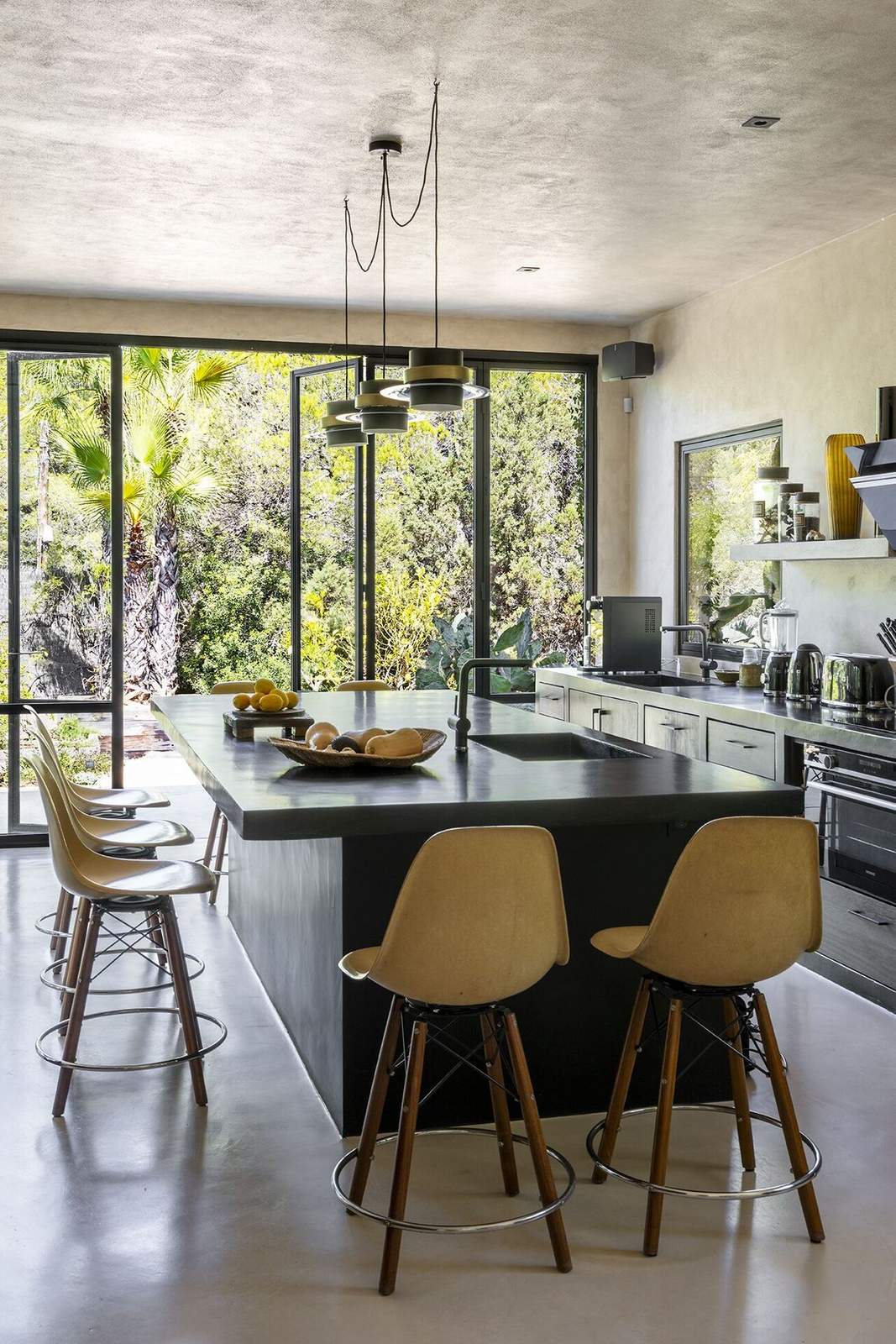 Black concrete kitchen island with vintage Eames bar stools and palms beyond