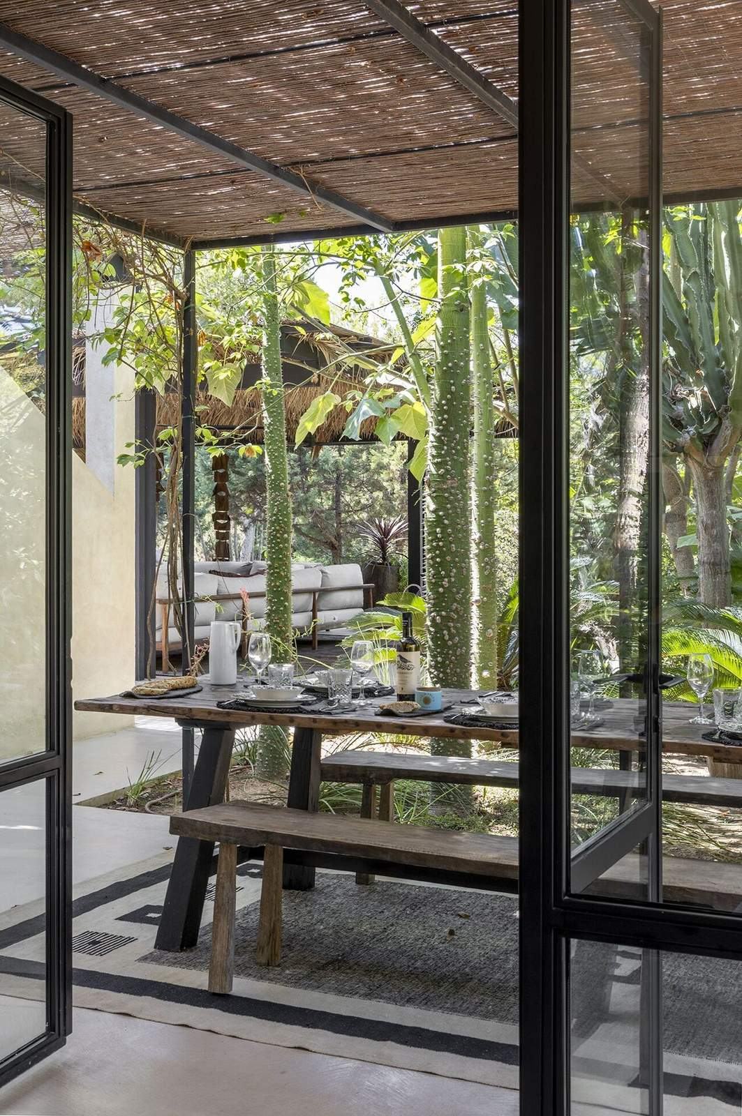 Looking through steel-framed doors to a wooden outdoor dining table under a reed pergola