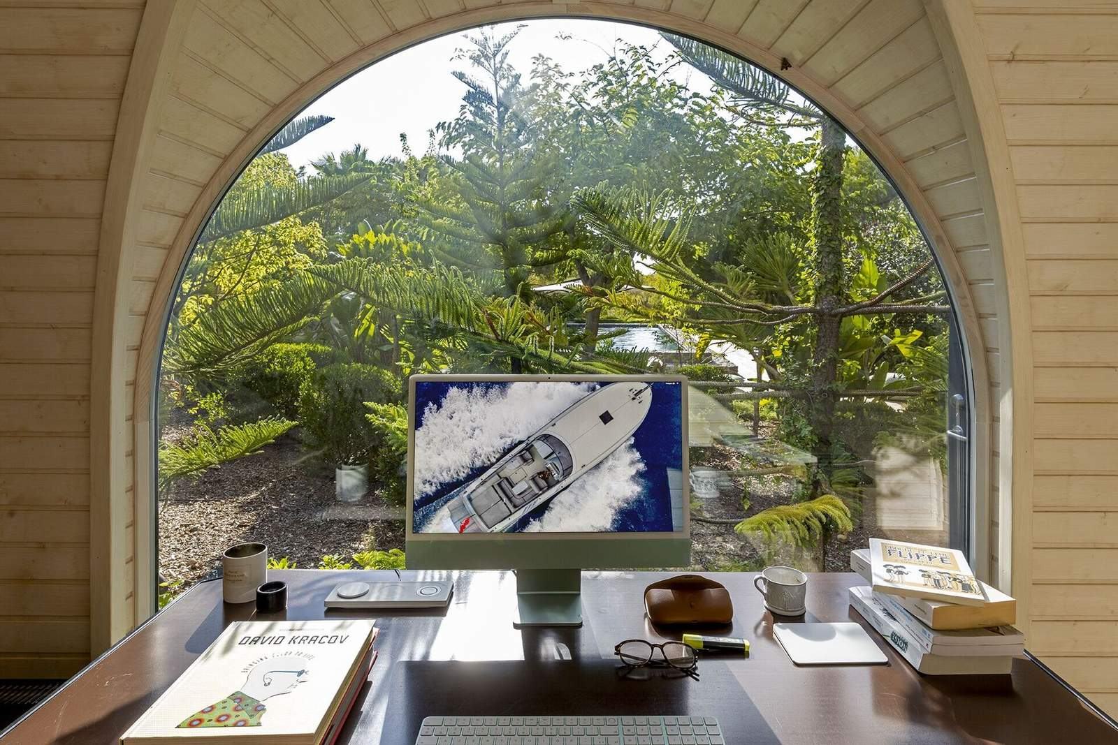 Office desk with iMac and books beneath the arched window looking out to the pool and garden