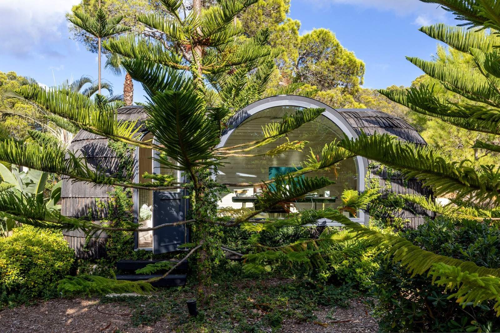 Front view of the shingled igloo office with arched window and door framed by ferns and pines