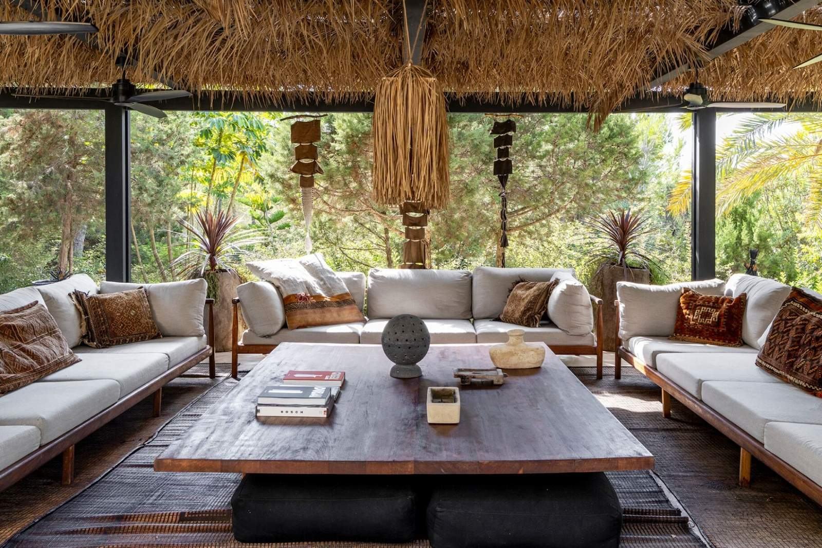 Balinese-style thatched pergola with three deep linen sofas around a hardwood coffee table, opening to a pine garden