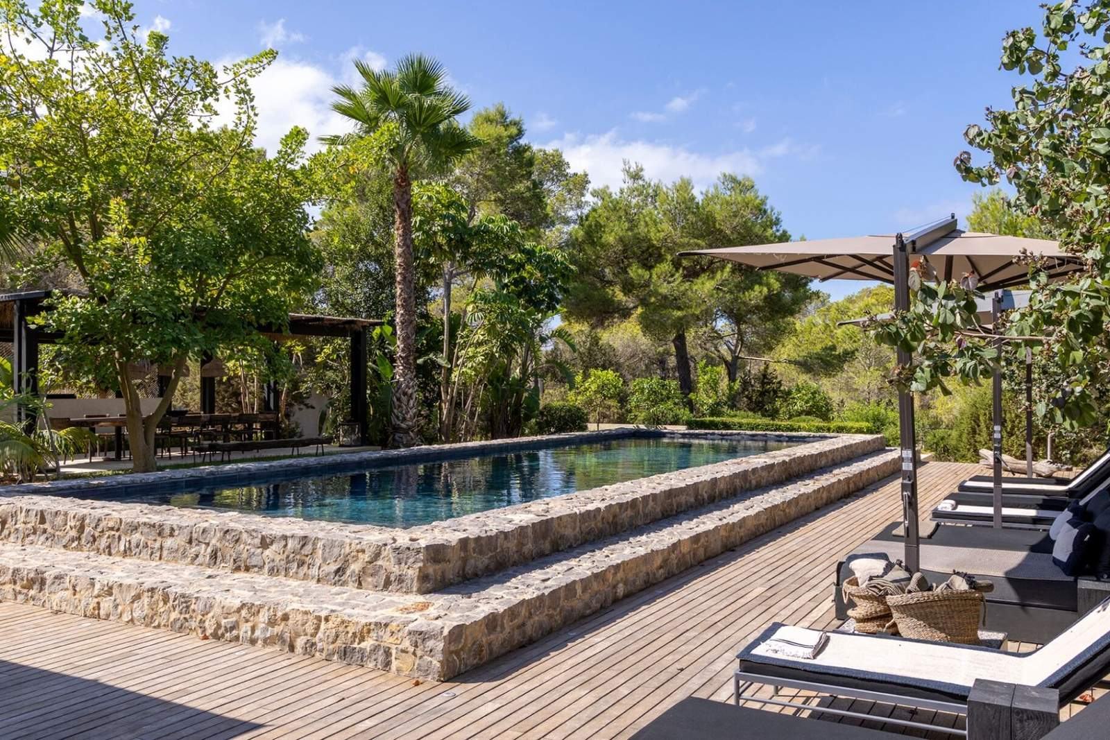 Stone-edged pool with teak deck, loungers and parasol under tall pines
