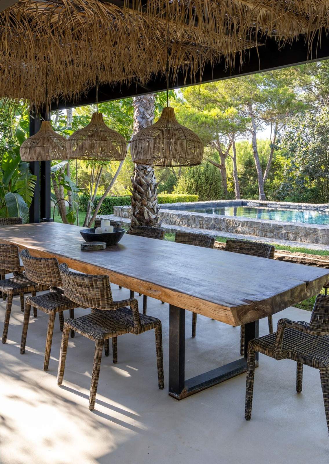 Live-edge dining table under woven lanterns with the stone-edged pool beyond