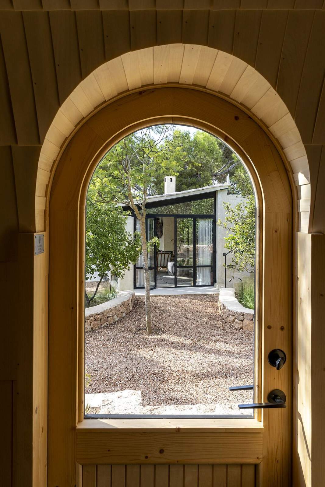 Arched timber sauna door framing a view of the gravel garden and main house