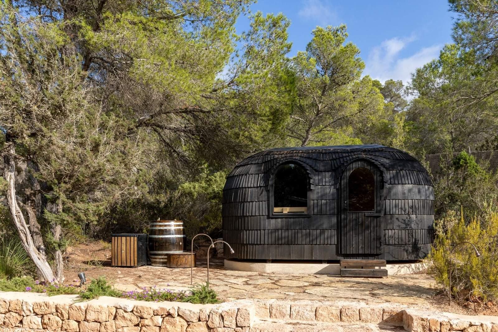 Black-shingled domed sauna pavilion among pines with ice plunge barrels alongside